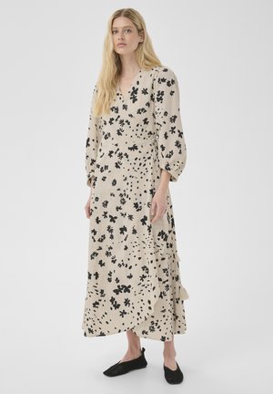 Mujer rubia con vestido cruzado beige con estampado floral negro y mangas tres cuartos abullonadas, de pie frente a un fondo claro y liso.