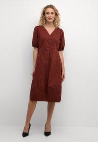 Rudo spalvos suknelė su V formos iškirpte, pūstomis trumpomis rankovėmis ir A linijos siluetu. Pagaminta iš lygios medžiagos, ji turi kelių ilgio kirpimą ir be raštų.