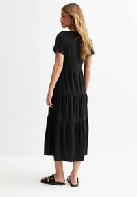 Vestido maxi negro de varias capas con mangas cortas, cuello redondo y tejido suave. Presenta una falda fluida y se lleva con sandalias negras.