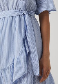 Gestreiftes Kleid in Blau und Weiß mit gebundenem Taillengürtel, das ein fließendes Design, einen ruffled Saum und einen strukturierten, leichten Stoff aufweist.