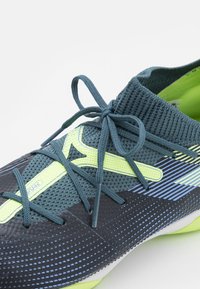 Scarpa sportiva nera e teal con tomaia in maglia testurizzata, dettagli giallo neon, lacci sottili e un design elegante e a profilo basso.