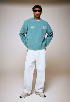 Čovjek u tirkiznom džemperu s logotipima, bijelim hlačama, tenisicama i crnim sunčanim naočalama stoji s rukama na leđima ispred jednobojne pozadine.