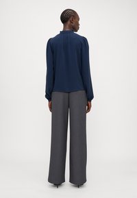 Blusa blu navy con scollo arricciato e maniche lunghe, abbinata a pantaloni grigi a vita alta e gamba larga, caratterizzati da un tessuto testurizzato.