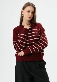 Bordeaux rode gebreide trui met witte horizontale strepen, voorzien van een geribbelde zoom en manchetten. Zachte textuur, losse pasvorm en ronde halslijn.
