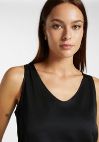 Top nero senza maniche con scollatura a V profonda. Tessuto liscio e lucido con cuciture minime. La modella ha un tatuaggio sul braccio sinistro.