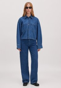 Denim-Outfit mit einer kurz geschnittenen Knopfleistejacke und locker sitzenden Hosen. Beide sind dunkelblau mit einer dezenten gemusterten Textur. Schwarze Sonnenbrille.