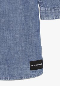 Svetle modrá džínová košile s tkanou texturou, která má na okraji černou nálepku s nápisem "CALVIN KLEIN JEANS" v bílém textu.