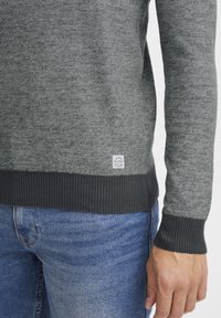 Gray knit sweater with a textured pattern, ribbed cuffs and hem in darker gray. Small brand label at the lower left. Worn over blue denim jeans.