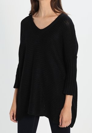 Kvinde iført løs sort strikket V-hals sweater med 3/4 ærmer, stående mod en ensartet lys baggrund.
