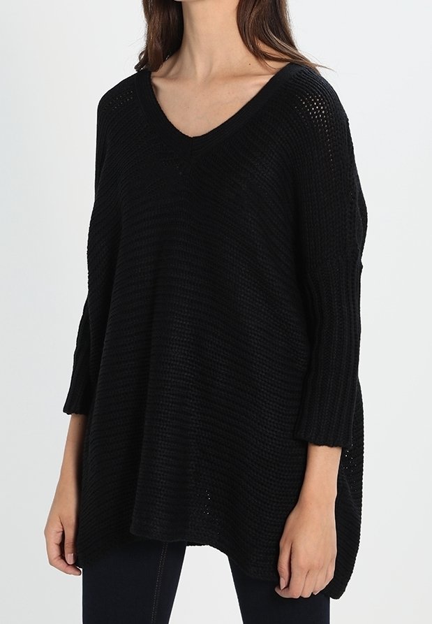 Femme portant un pull noir ample en maille avec un col en V et des manches 3/4, debout devant un fond clair uni.