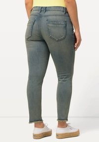 Úzké džíny z vybledlého šedého denimu, s elastickým pasem ve střední výšce, dvěma zadními kapsami a třásněmi na nohavicích. Nosené s bílými espadrilkami.