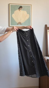 Falda de lunares en blanco y negro, de largo hasta la rodilla, que presenta una fila de botones en la parte delantera y una textura fluida. Sostenida contra un fondo neutro.
