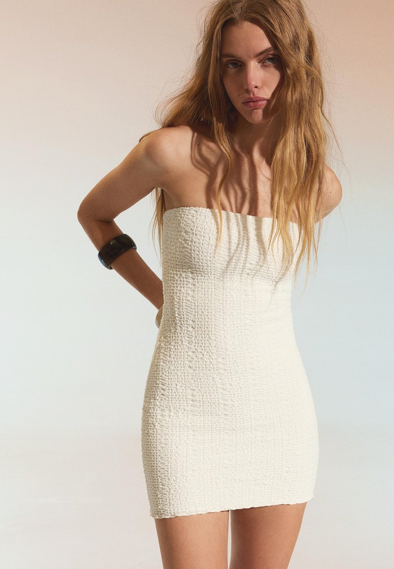 Frau mit langen Haaren trägt ein trägerloses, strukturiertes weißes Minikleid und ein schwarzes Armband, steht mit hinter dem Rücken verschränkten Armen.