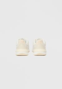 Ζευγάρι μπεζ και λευκά αθλητικά παπούτσια εμφανιζόμενο από την πίσω πλευρά σε απλό ανοιχτό φόντο, τονίζοντας τις λεπτομέρειες της φτέρνας και της σόλας.