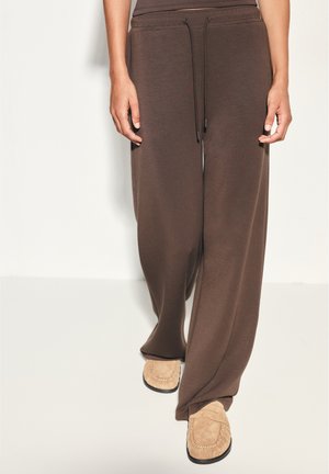 Personne portant un pantalon marron ample avec cordon de serrage et des mocassins beige clair marchant sur un sol clair devant un mur uni.