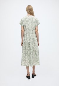 Femme aux longs cheveux blonds portant une robe mi-longue vert sauge à motif floral blanc et des sandales à talons noires, debout de dos.