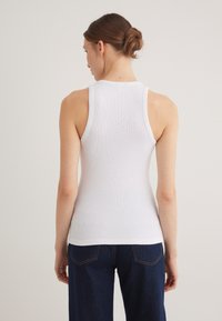 White ribbed tank top with a fitted design and wide straps, seen from the back. Paired with dark blue jeans. Smooth texture.
