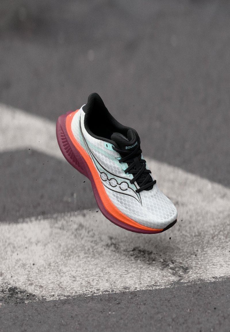 Hvit og svart løpesko med oransje og lilla såle svever over asfalt med hvit pilmerking.