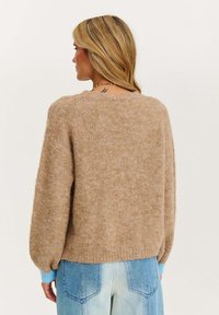 Beżowy sweter dziany o luźnym fasonie, z prążkowanym dołem i długimi rękawami, które mają jasnoniebieskie mankiety. Miękka faktura. Noszony z jasnymi dżinsami.