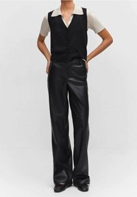 Personne portant une chemise beige à manches courtes avec col sous un gilet noir sans manches, associée à un pantalon large en cuir noir taille haute.