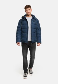 Giacca imbottita blu navy con felpa grigia sotto. Presenta una chiusura a zip e cappuccio con cordino, indossata con jeans grigi e sneakers multicolori.