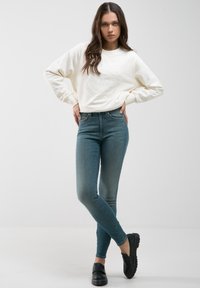 Balta džemperis su iškilniu 1979 metų detalimu, derinamas su prigludusiomis mėlynomis džinsomis ir juodais platforminiais batais. Lygūs paviršiai, kasdienis dizainas.