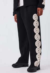 Schwarze Jogginghosen mit einem strukturierten cremefarbenen Häkelmuster an der Seite, aus weichem Stoff und in lockerer Passform.