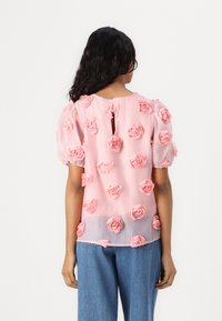 Top trasparente rosa con applicazioni floreali in rilievo, scollatura rotonda, schiena a chiave e maniche a sbuffo. Indossato con jeans in denim azzurro chiaro.