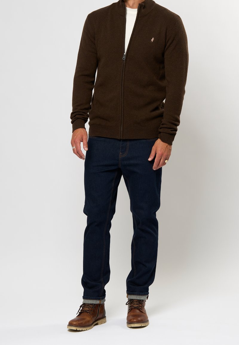 Homme portant un pull à fermeture éclair marron foncé, un jean bleu foncé avec revers, et des bottes en cuir marron à lacets, debout sur fond blanc.