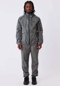 Giacca impermeabile grigia con pantaloni abbinati. Caratterizzata da una chiusura lampo frontale, cappuccio regolabile e dettagli neri. Texture liscia, vestibilità atletica.