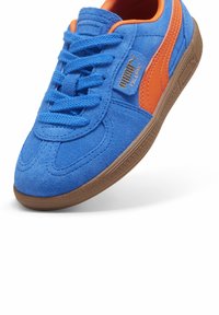 Mėlynos zomšinės sportbačiai su oranžiniais akcentais, plokščiais raišteliais, rudos gumos padu ir auksine ženklodara šone.