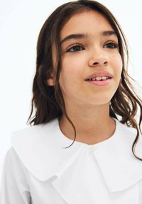 Valkoinen kauluspaita, jossa on suuri, litteä kaulus ja nappisulku kaulan kohdalla. Suorasilkkiinen kangas, jota käyttää lapsi, jolla on löysät, laineikkaat hiukset.