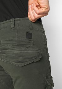 Oliwkowe spodnie cargo o wąskim kroju, z wieloma kieszeniami, teksturowaną tkaniną oraz czarną naszywką z logo z tyłu.