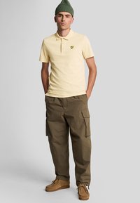Lichtgeel polo-shirt met een klein zwart logo, gecombineerd met losse olijfgroene cargo-broeken en beige suède schoenen.