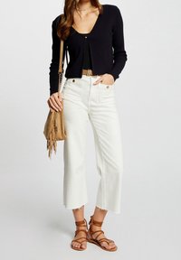 Cárdigan negro acanalado con escote en V, combinado con jeans blancos recortados de cintura alta. Un bolso de flecos marrón y sandalias marrones con tiras completan el look.