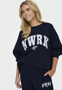 Felpa blu navy con "NWRK" stampato in bianco. Presenta una vestibilità ampia, un colletto rotondo e una texture morbida. Abbinata a pantaloni blu navy coordinati.