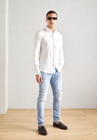 Hvit lin skjorte med knapper, lyseblå slitte bukser og svarte loafers. Modellen har på seg svarte rektangulære solbriller, står i en minimalistisk setting.