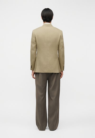 Άτομο με κοντά μαύρα μαλλιά που φοράει μπεζ σακάκι και άνετο καφέ παντελόνι, στέκεται γυρισμένο με την πλάτη προς ένα απλό φωτεινό φόντο.