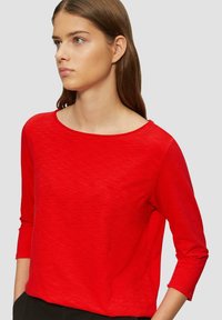 Haut rouge à manches longues avec un col rond, fait de tissu doux et texturé. Présente une coupe décontractée et des manches de longueur intermédiaire.