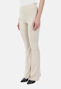 Donna che indossa pantaloni beige a gamba flare aderenti abbinati a tacchi a punta neri, in piedi davanti a uno sfondo bianco.