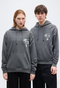 Δύο νεαροί ενήλικες φορούν πανομοιότυπες γκρι φούτερ με κουκούλα και λευκό κε ίμενο στο στήθος, στέκονται μπροστά σε λευκό φόντο.