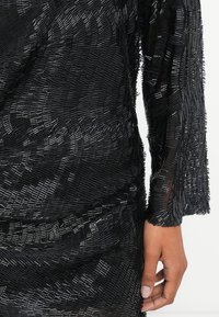 Melns teksturēts apģērbs ar slāņainu pērlīšu apdari, ar garām piedurknēm un pieguļošu piegriezumu, uzsverot sarežģītus rakstus un dziļumu.