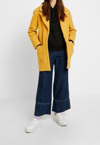 Cappotto giallo texture con ampi rever, felpa nera sotto, pantaloni in denim larghi scuri e sneakers bianche con chiusura a lacci.