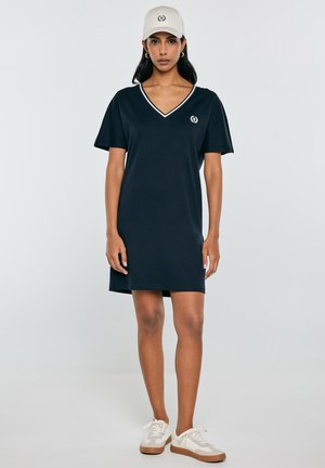 Kobieta ubrana w granatową sukienkę z krótkim rękawem i dekoltem w serek, beżową czapkę z logo oraz białe trampki, stojąca na jednolitym jasnym tle.