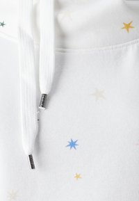 Sudadera blanca con patrones de estrellas dispersas en azul, verde, amarillo y beige. Cuenta con cordones con extremos metálicos. Textura suave.