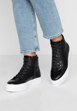 Czarne sneakersy typu high-top z teksturowanym wzorem, białe grube podeszwy i odblaskowe sznurowadła, noszone z jasnoniebieskimi skróconymi dżinsami.