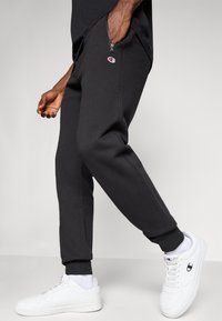 Mand iført sorte sweatpants med lynlås-lommer og hvide sneakers med Champion-logo, mens han går mod en ensfarvet baggrund.
