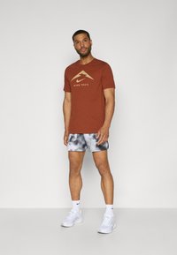 Brun T-skjorte med gull Nike Trail-logo, kombinert med grå tie-dye shorts. Hvite treningssko og sokker fullfører antrekket.