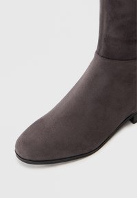 Close-up de uma bota de camurça cinza escuro com ponta arredondada e salto baixo preto sobre um fundo branco.