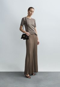 Brązowa prążkowana koszulka w połączeniu z brązową spódnicą o szerokich nogawkach. Modelka trzyma małą czarną torebkę na ramię. Gładka tekstura, minimalistyczny design.